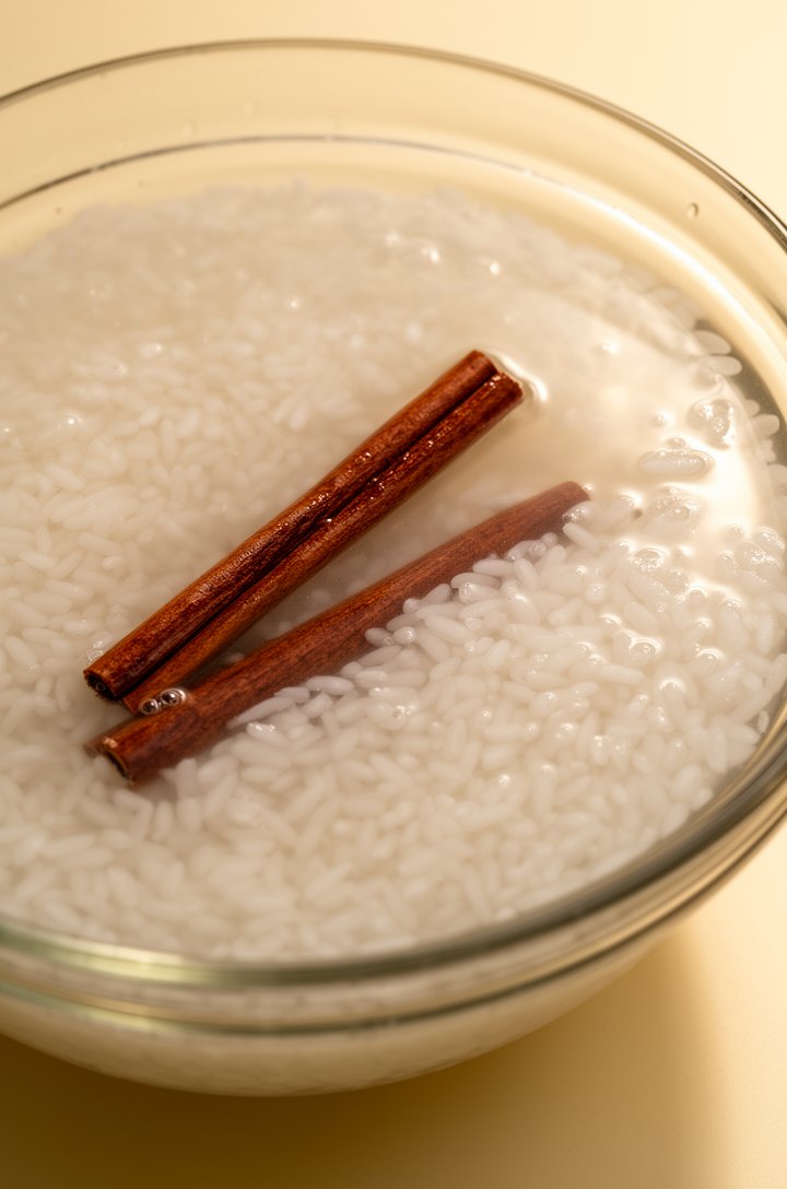 Close-up 45-degree angle of a glass bowl filled with white rice soaking in cloudy water with two cinnamon sticks submerged, rice grains visibly swollen, warm side light catching the water surface, but