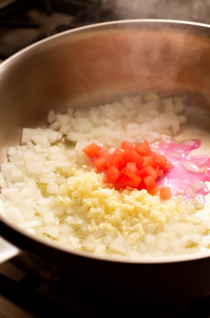Close-up 30-degree angle of a large saucepan on a stove, translucent diced white onion and minced garlic sizzling gently in melted butter, tiny pieces of fresh tomato just added and releasing pink jui